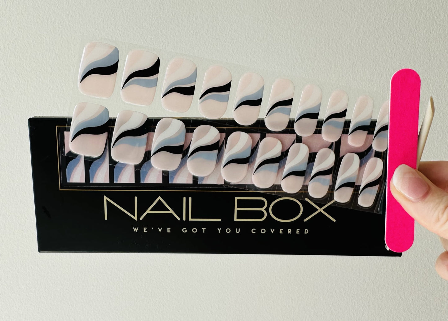 a female hand holding nail file and orange stick and wave good by a semi cured gel nail sticker set which is pink with blue black and white wave across the center sold by Nail Box